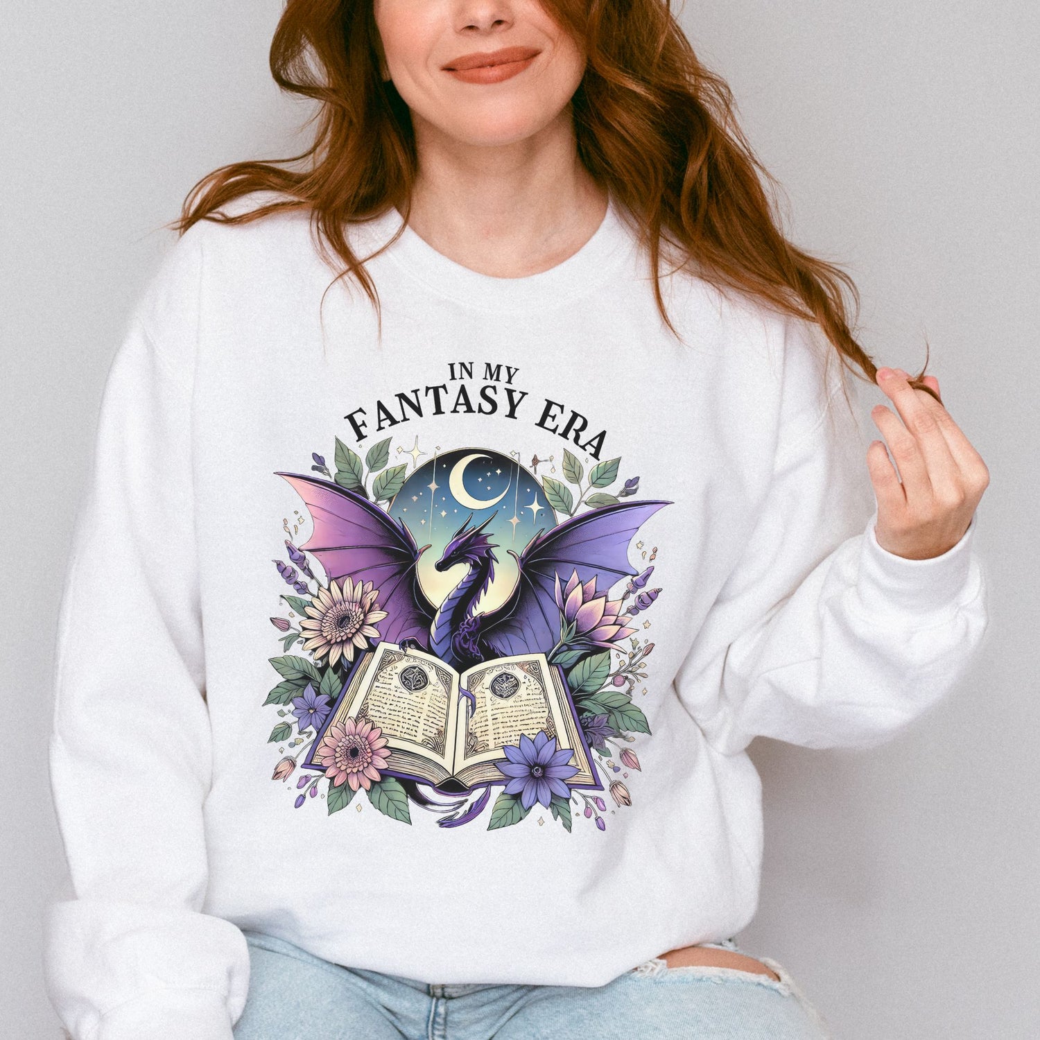 The in my fantasy era sweatshirt has a design of a purple dragon behind an open fantasy book and they are surrounded by purple and pink flowers. The text above the dragon says: in my fantasy era.
The model is wearing the colour white.